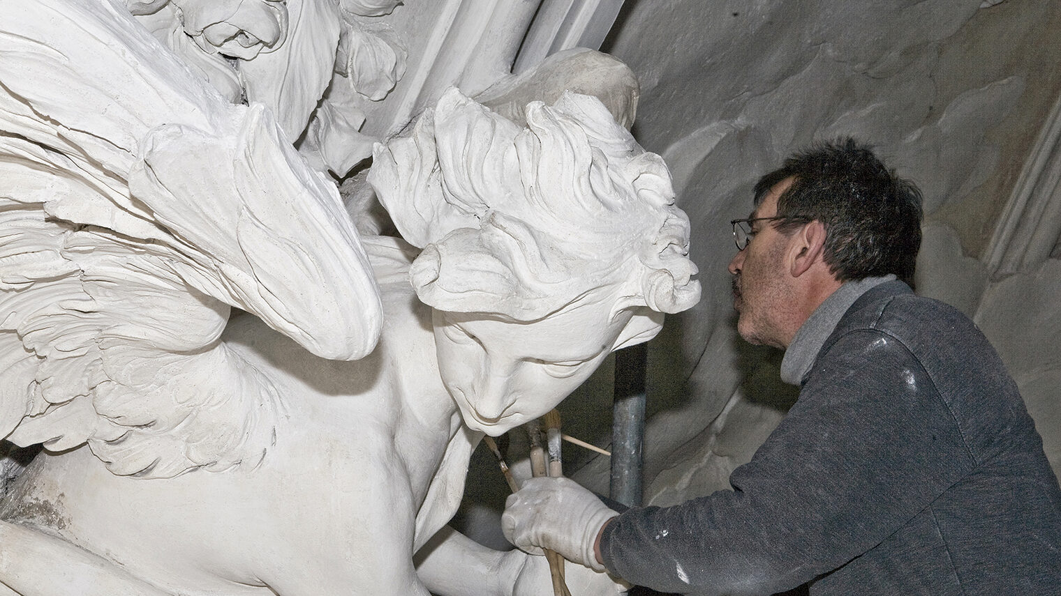 Auf dem Bild ist ein Restaurator bei der Konservierung und Restaurierung von Stuckelementen an der Apsis der Kollegienkirche Salzburg zu sehen.